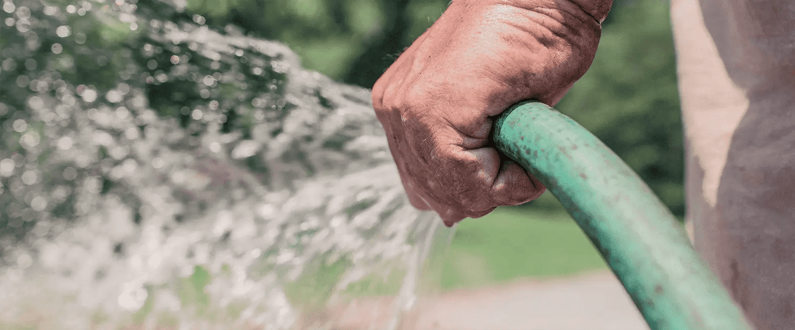 Nahaufnahme einer Hand, die einen grünen Gartenschlauch hält, aus dem Wasser spritzt.